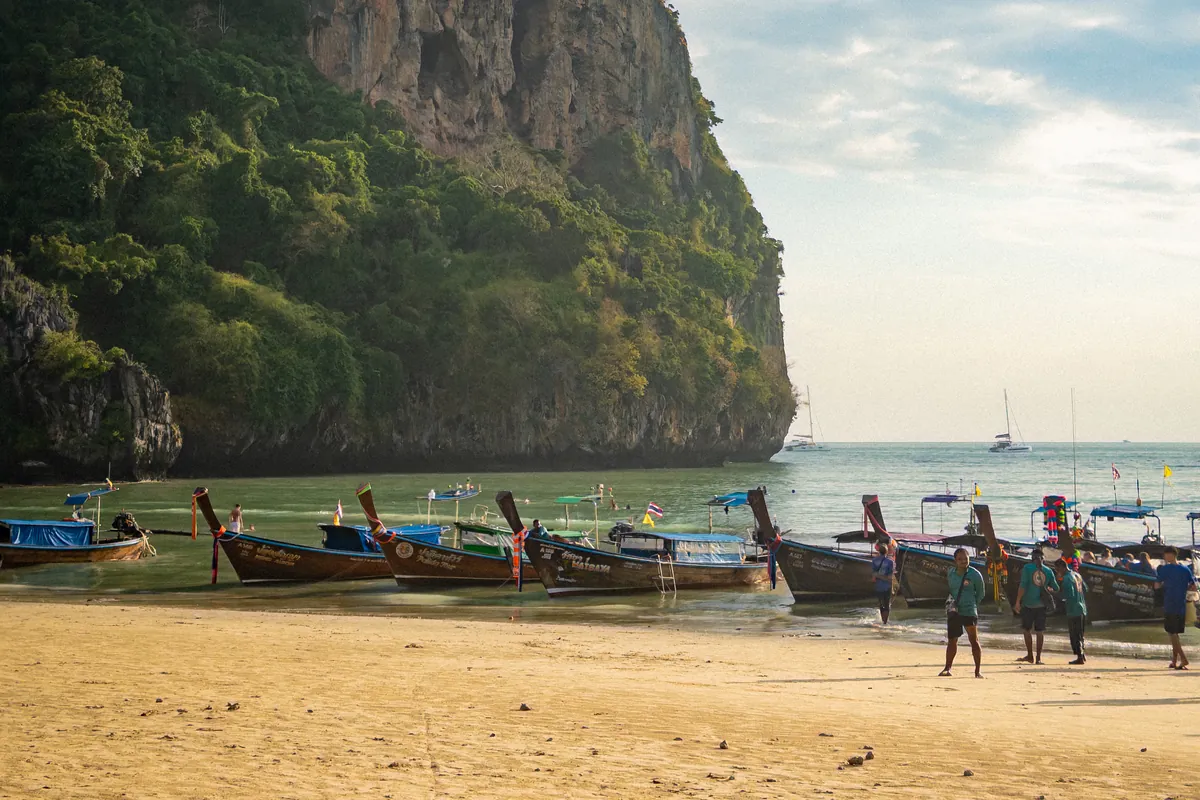 Longtail boats lined up on the sand at Railay West Beach beneath a tall limestone cliff