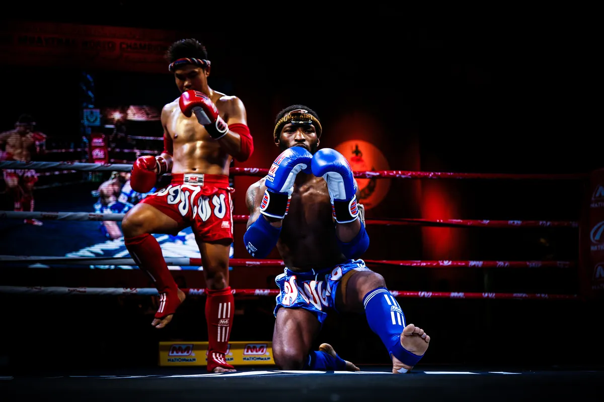 Two Muay Thai fighters performing the ram muay in a ring under stage lights, one on a raised knee and one kneeling in blue gloves