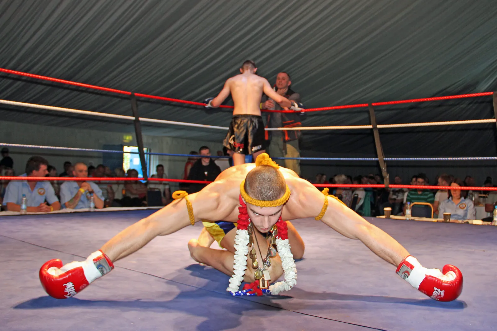 A Muay Thai fighter in a gold headband and armbands bowing low in the ring during the Wai Khru Ram Muay, with spectators at ringside